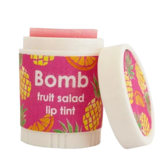 Lip tint container labeled "Bomb" featuring a playful design with pineapples and orange segments on a pink background, with the text "Bomb fruit salad lip tint" displayed prominently on the front. The cap is slightly open, revealing the pink tint inside.